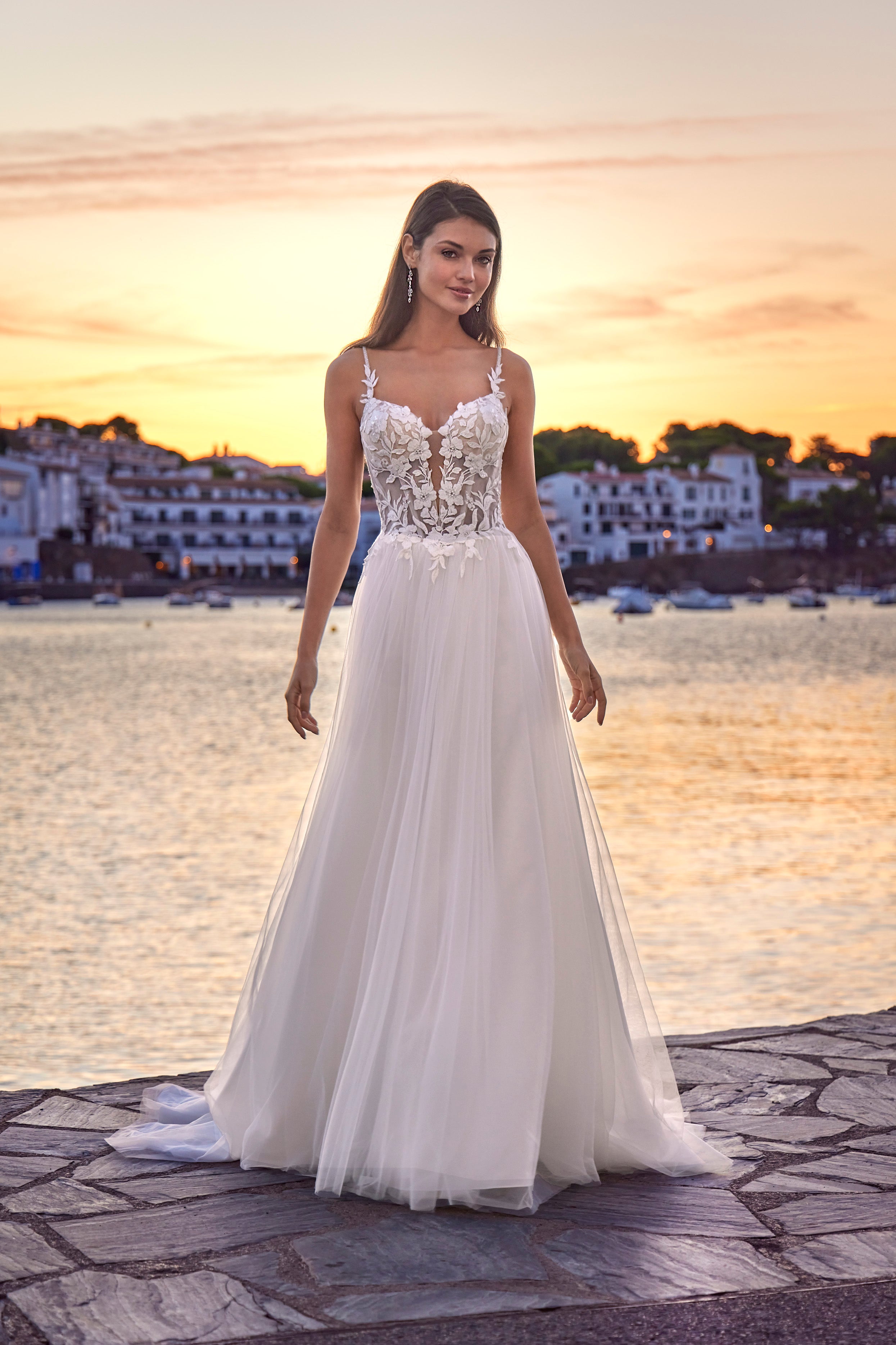 Woman in a white wedding dress standing by a waterfront at sunset.