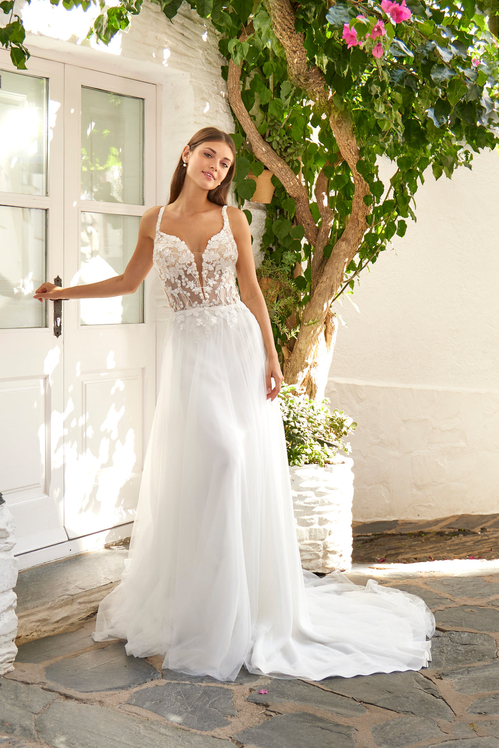 Woman in a white wedding dress standing outdoors with greenery and a building in the background
