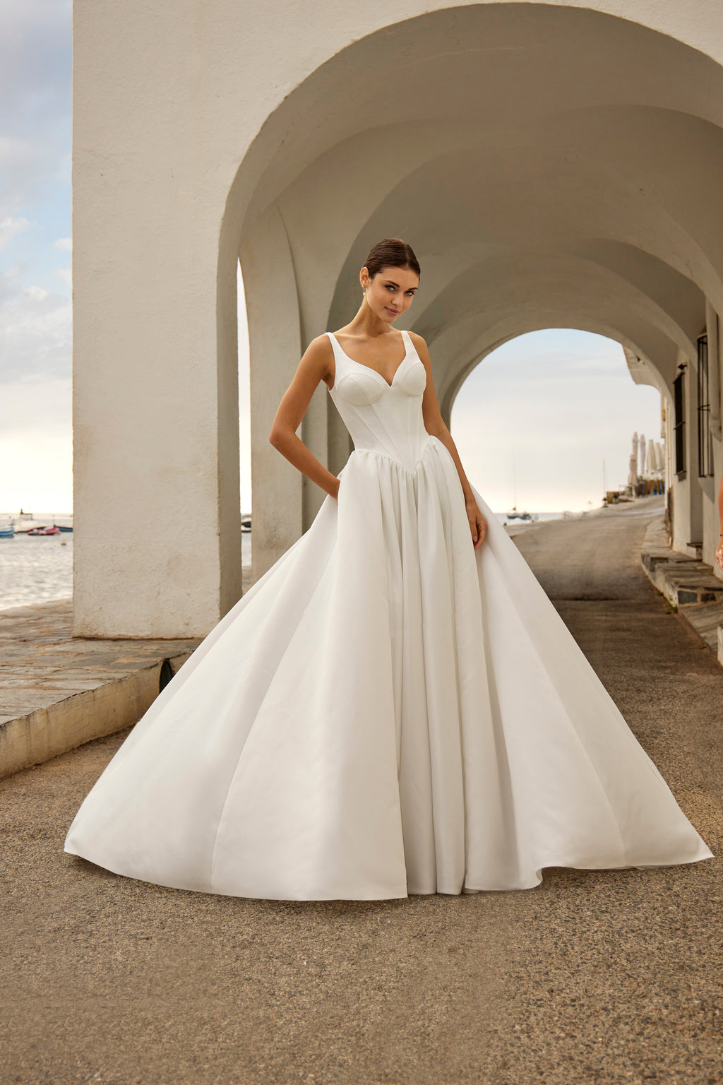 Woman in a white wedding dress standing under a white archway with a coastal background.