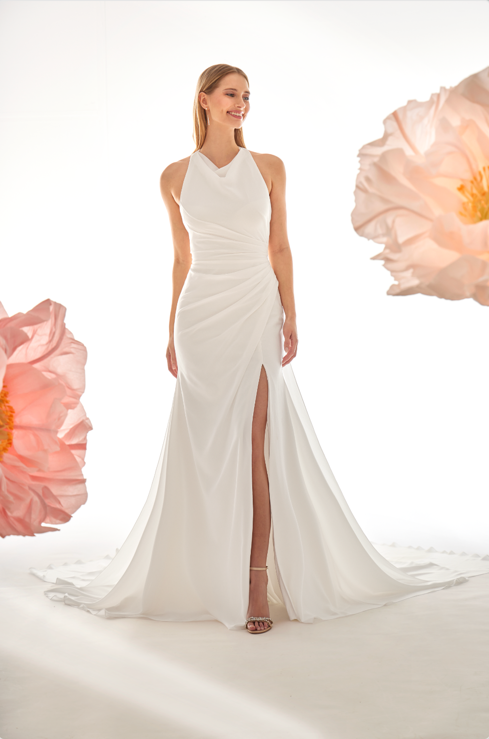 Woman wearing a white halter neck gown with a high slit, surrounded by pink flowers on a light background.